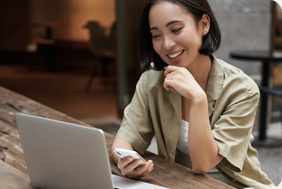 A woman smiling at her phone while shopping on a laptop